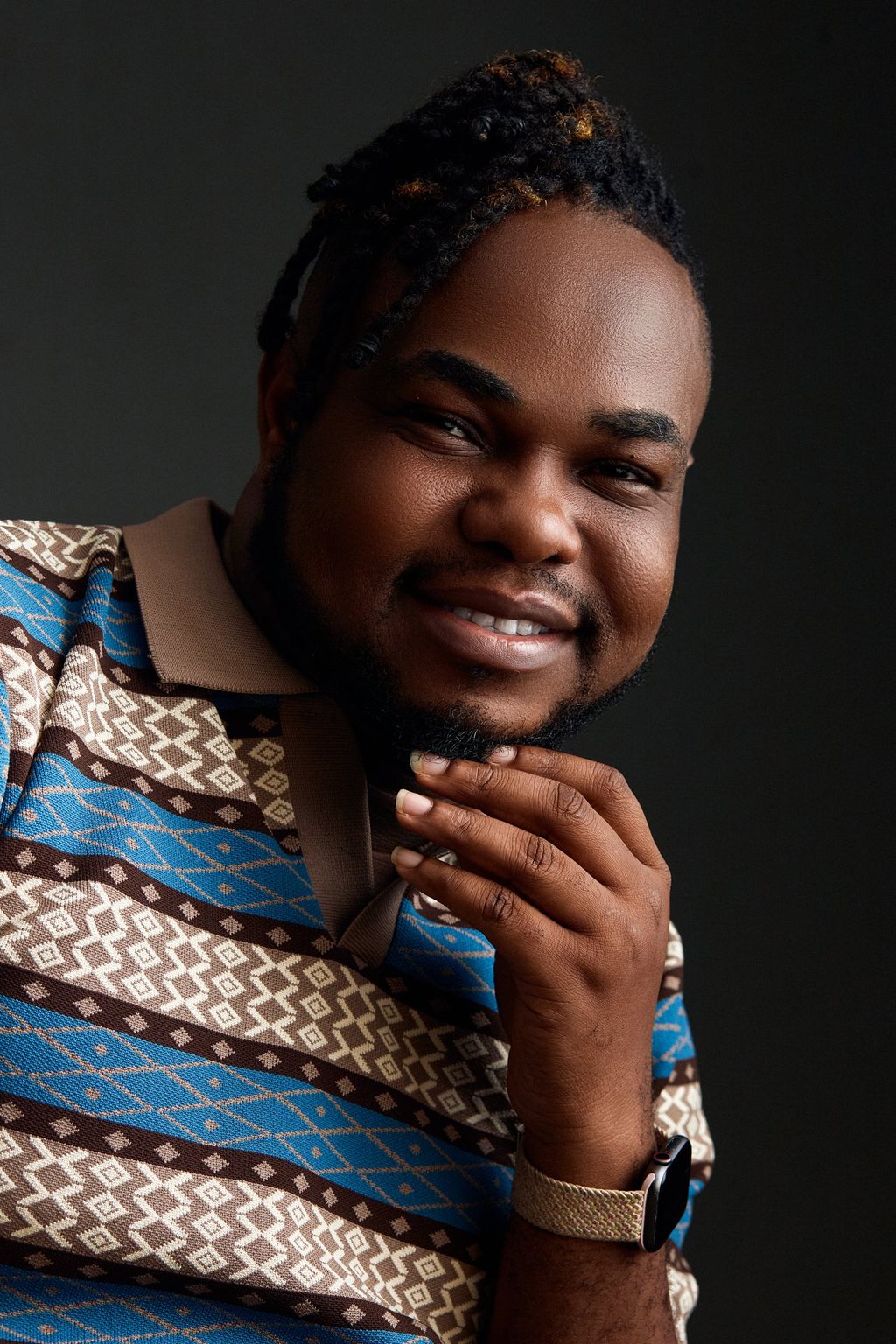 Tai Ajayi, smiling, chin resting on his hand, in a blue and brown patterned shirt