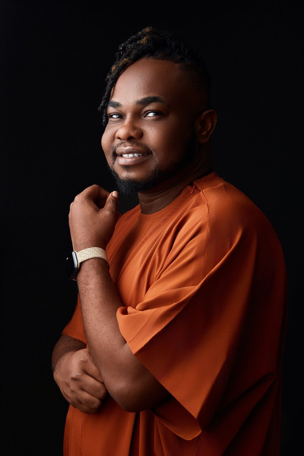 Tai Ajayi in an orange kaftan, smiling, hand raised to his chin